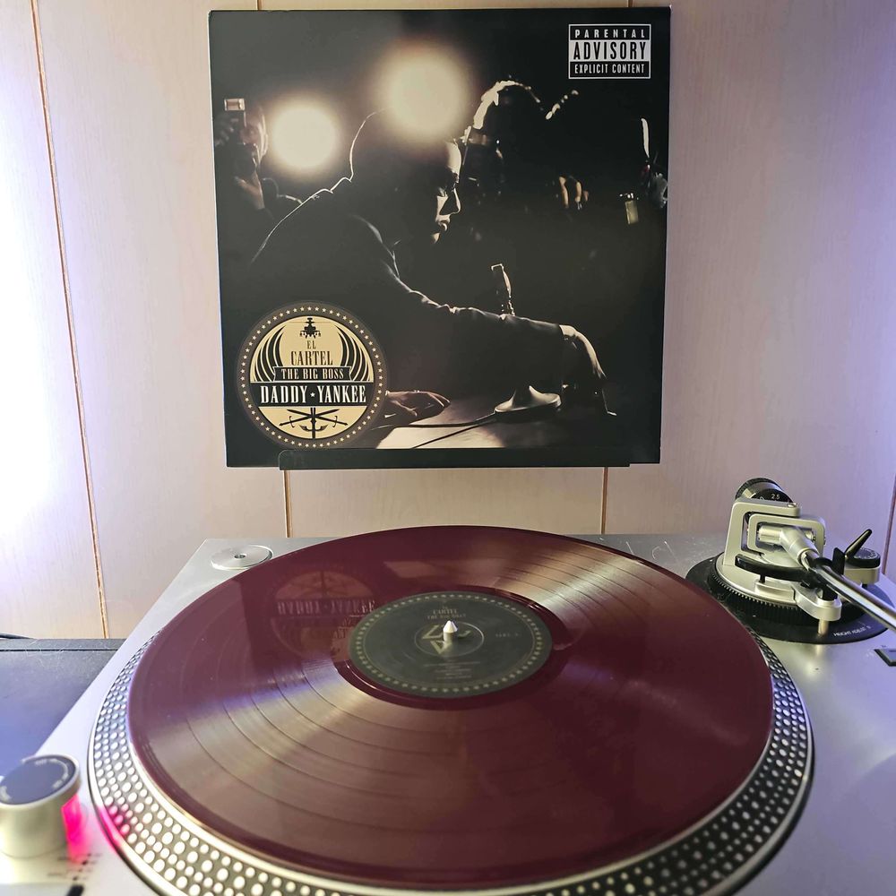 A Translucent Purple vinyl record sits on a turntable. Behind the turntable, a vinyl album outer sleeve is displayed. The front cover shows Daddy Yankee sitting at a table with a mic in front of him. In the background are a bunch of photographers