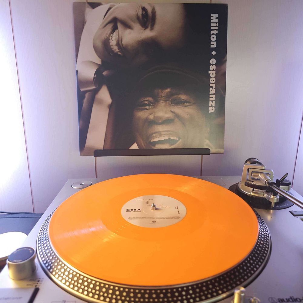 An orange vinyl record sits on a turntable. Behind the turntable, a vinyl album outer sleeve is displayed. The front cover shows Esperanza Spalding's and Milton Nascimento's faces close together as they smile at the camera.