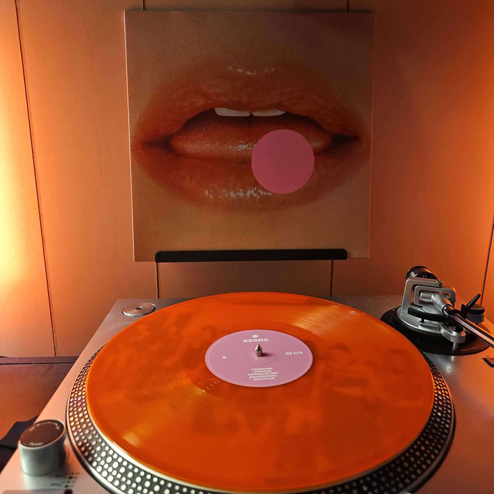 An orange vinyl record sits on a turntable. Behind the turntable, a vinyl album outer sleeve is displayed. The front cover shows Kesha's mouth and pink dot covering part of it.