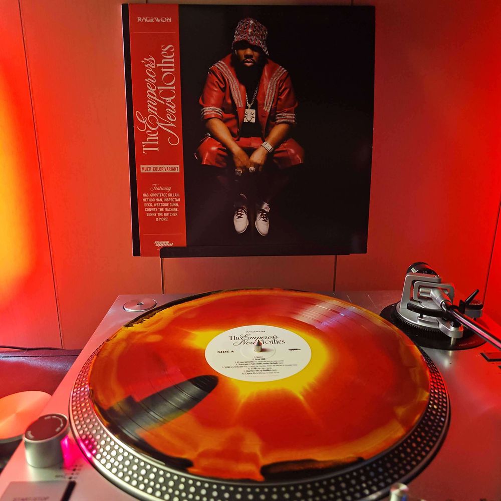 A Red,Yellow & Black Mix (Ocean Cranfire) vinyl record sits on a turntable. Behind the turntable, a vinyl album outer sleeve is displayed. The front cover shows Raekwon sitting down. He's wearing red leather jacket and short, and a large chain with a spider medallion.