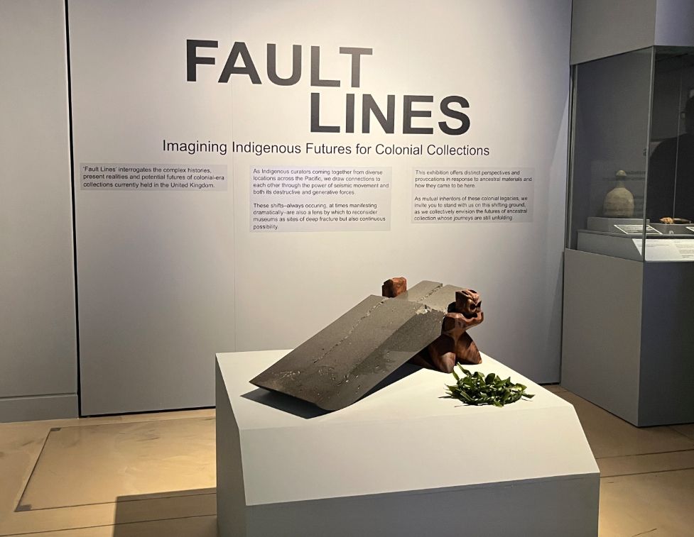 A contemporary sculpture featuring a rectangular-shaped split slab of stone which is held together by two carved wooden figures. On the wall behind the sculpture is text which includes the exhibition's title 'Fault Lines: Imagining Indigenous Futures for Colonial Collections'.