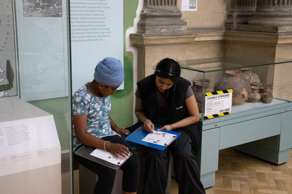Two people sit in a gallery with clipboards. They are colouring in pictures of objects on display in the museum.