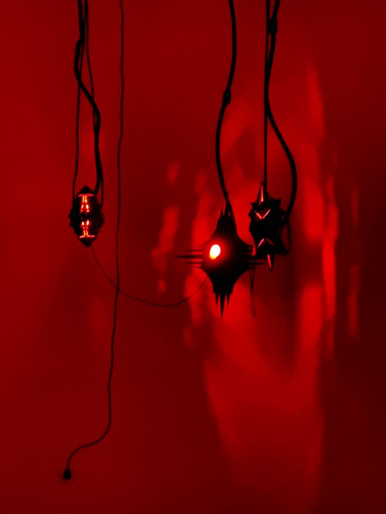 3-D printed sculptures by Embryoroom hanging suspended from black wires. The wall behind is lit in red and the the objects cast eerie shadows.