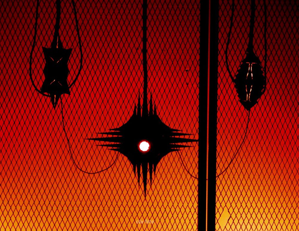 A red lit view of three hanging sculptural objects viewed from behind a cage. In front of them is the mesh of a metal cage. Wires hang from and connect each object. They are lit from within and have alien, futuristic shapes. The central one has red eye shining from its centre. The top, bottom and sides of the black object end in five sinister spikes.