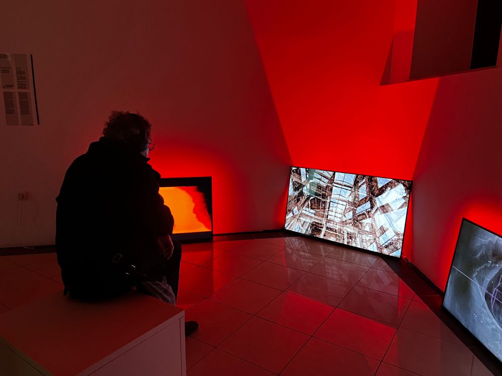 A viewer at the exhibition T.F.I.D. (The Future is Dead) sits watching and listening to the video "Degeneracija [TEV-24]" by Sz. Berlin ± Panic. The screen shows the interior of a vast industrial space. A red beam illuminates the wall behind the screen. 2 other screens are visible to the left and right. 