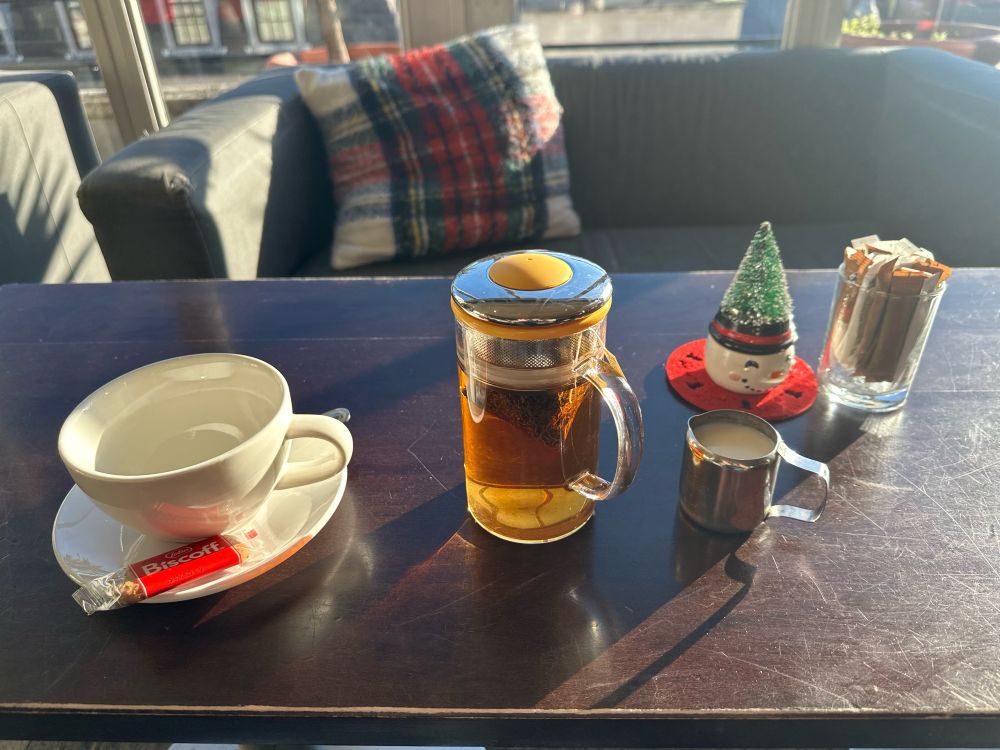 An empty cup, a tea pot brewing tea, a small jug of milk, a Christmas table ornament and a small glass with sugar sachets on a table in the sunshine. 