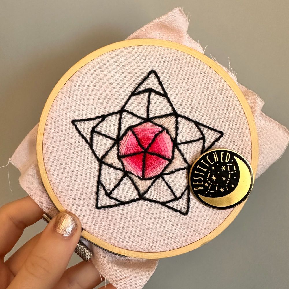 math art/ mathematical embroidery of a star from the Penrose tiling in an embroidery hoop. It is a black outline of a star with the kite middle sections filled in with a gradient pink, and the darts in the middle filled in with a baby pink. 