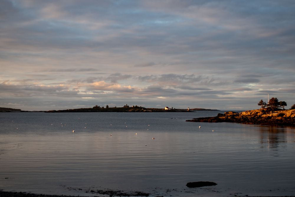 Ram Island, East Boothbay Maine, sunlight raking across buildings and mooring buoys. Blue sky and clouds in the distance.