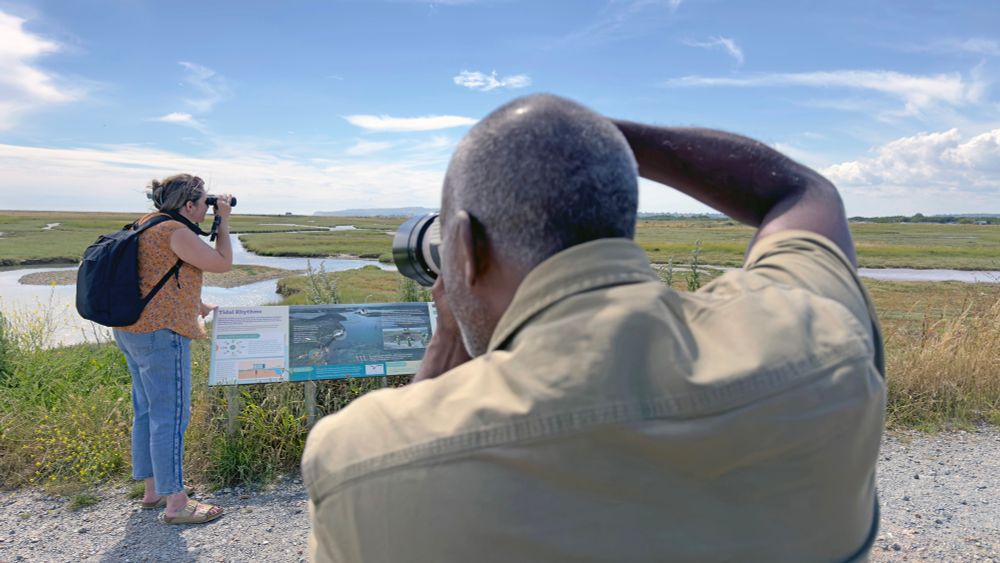Man filming woman looking through binoculars at nature reserve