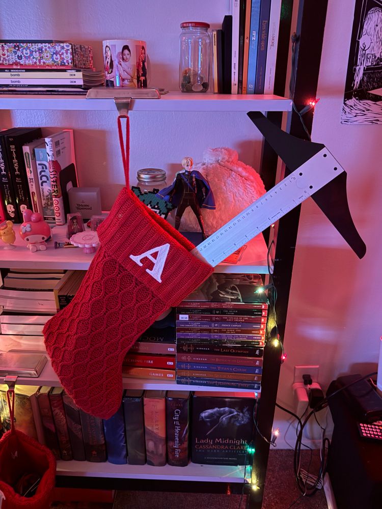 a red stocking hangs from a bookshelf full of books and trinkets. There is a white letter A on the knit fabric and a t-square sticks out of it very obviously. 
