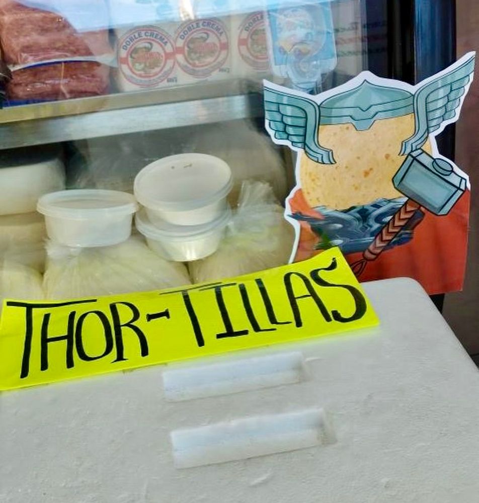 A yellow sign is placed prominently on a shop’s counter in front of the food. It's hand-lettered in black and reads: THOR-TILLAS. 