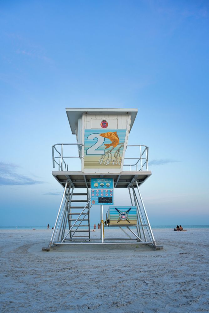 A lifeguard tower with the number 2 on it. People are in the back ground as is the Gulf of Mexico. An image of a fish is next to the tower number
