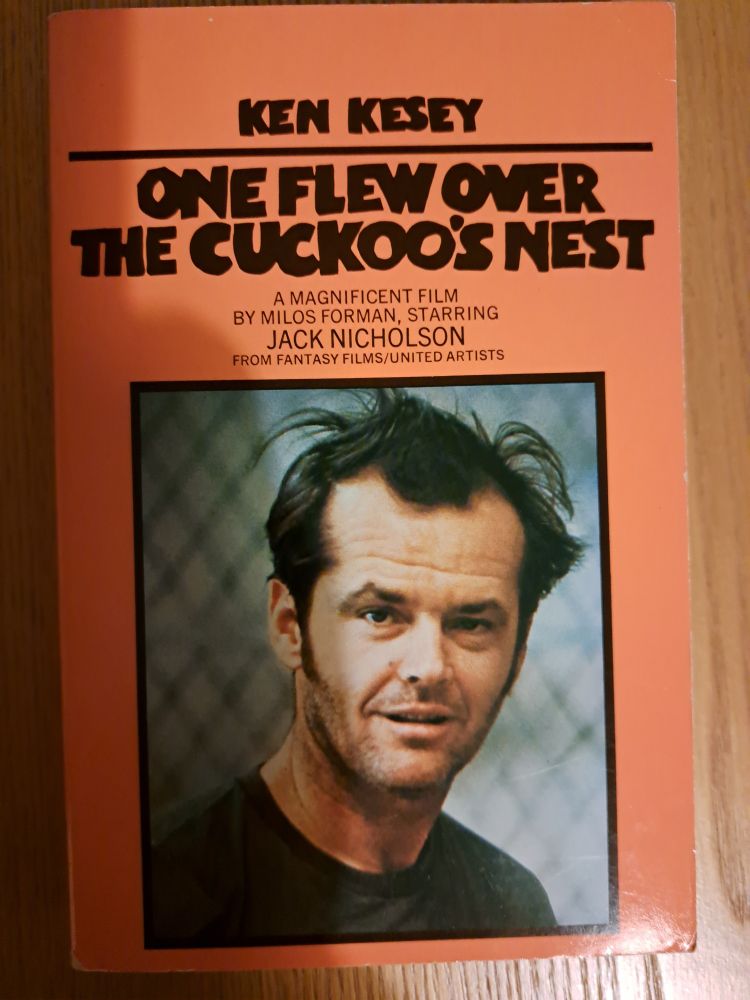 Front cover of the book 'One Flew Over The Cuckoo's Nest' by Ken Kesey. Shows an orange background and a picture of Jack Nicholson in his role in the film if the same name 