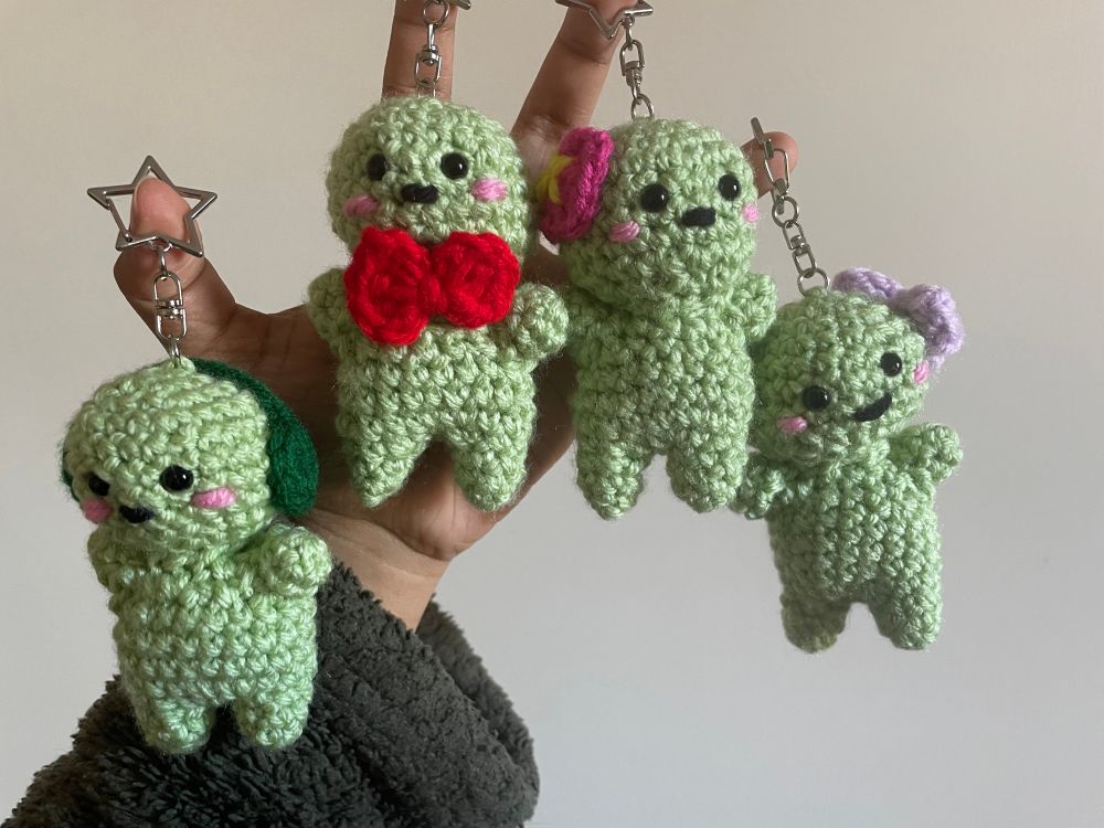 4 crochet smiski keychains. from left to right: smiski with green headphones, smiski with red bow tie. smiski with pink flower, smiski with purple bow