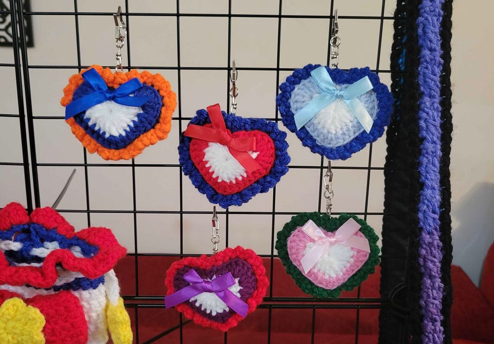 5 heart shaped crochet keychains inspired by the outfits of sailor scouts (sailor moon, mercury, mars, venus, and jupiter) with star shaped clasps. all the hearts have white centers and a frill border matching the color of the sailor scout’s skirt. keychains are hung on a black metal grid panel. 