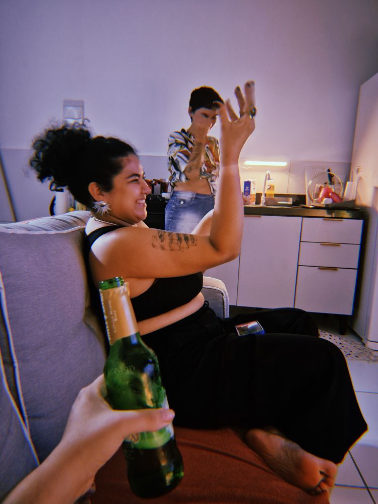 Raisa, bela também porem muito bem casada, com seu lookinho preto top calça noite despretensiosa, fazendo uma mãozinha italiana para a massa que estávamos prestes a saborear, sentada no sofá de bruna mulher que ao fundo ri fazendo o mesmo gesto com a mão enquanto aparece só minha mãozinha segurando uma cerveja