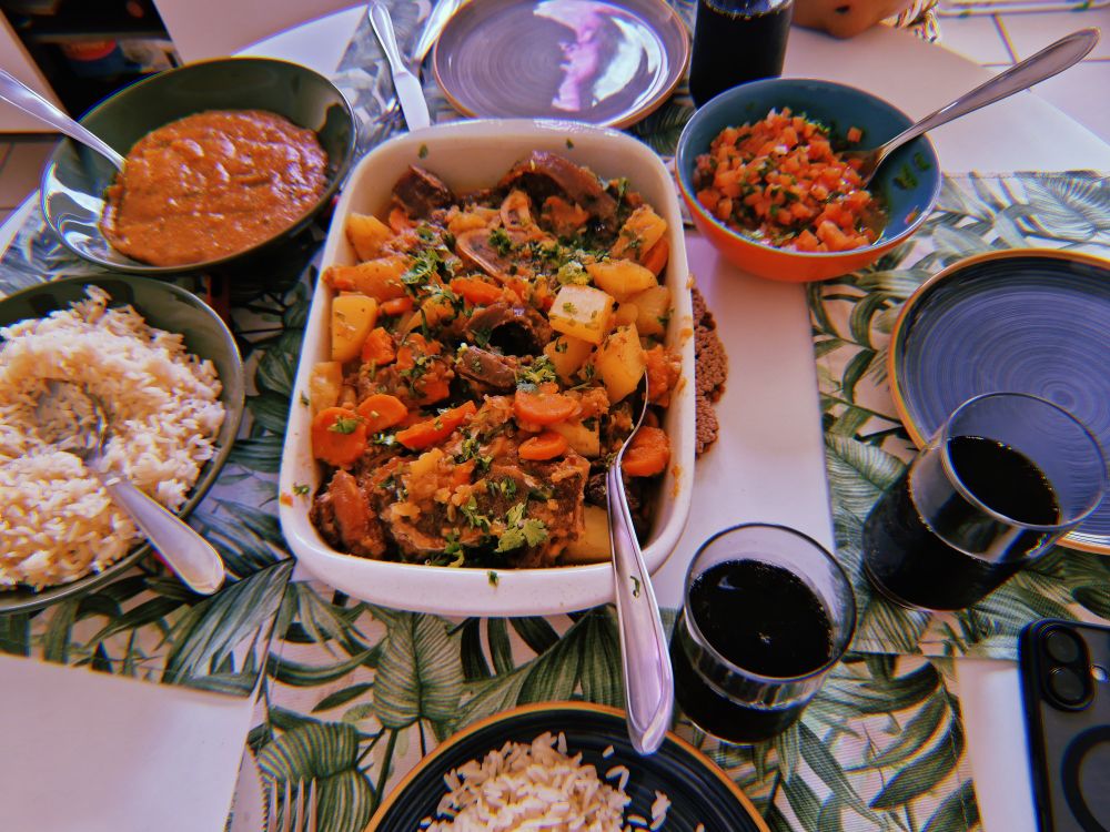 Fotografia de uma mesa farta com uma travessa de carne de mocotó, pirão, arroz, vinagrete e coquinha pra acompanhar, um jogo americano de folhas e pratos belissimos feitos por minha amiga lija