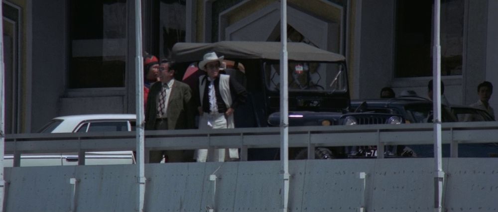 Shot from a distance. There's a couple of guys here. Middle aged guy in a suit and most importantly David Chiang wearing a black shirt, long sleeveless white coat, white hat and white trousers. Possibly sunglasses
