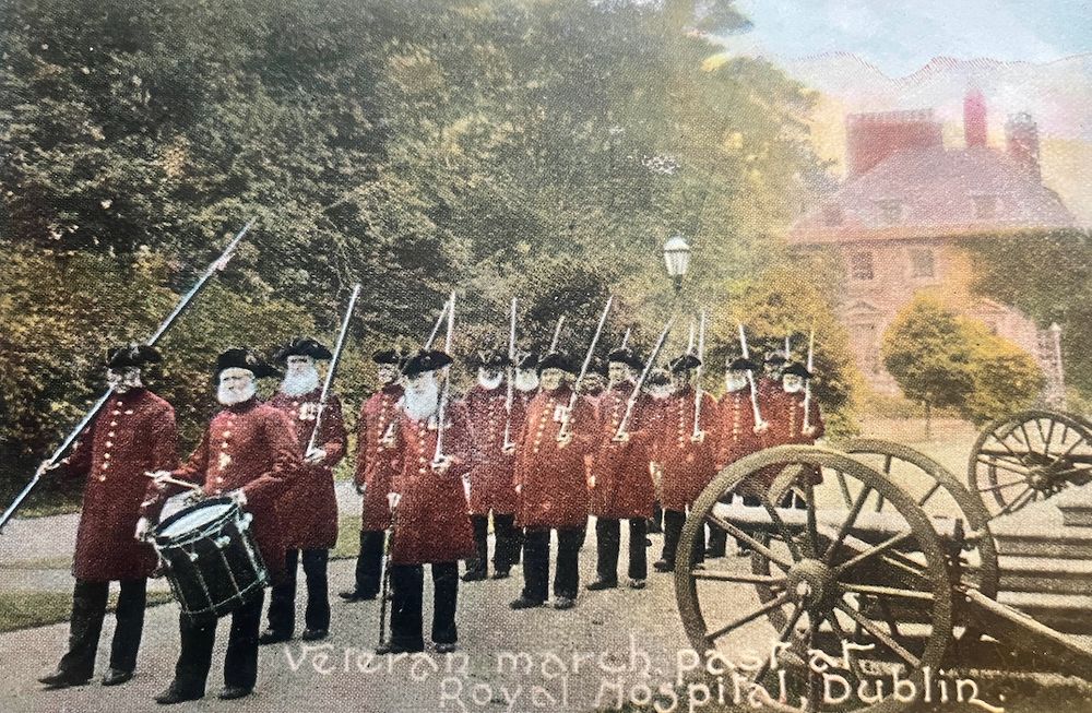 14 elderly men in the uniform of the Royal Hospital Kilmainham (red coats, tricorn hats) marching two abreast and carrying arms, the foremost carrying a drum.