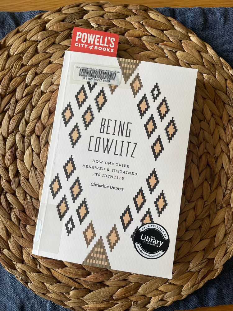 Paperback book atop a woven, grass, circular mat. The book has a cream colored cover with light and dark brown diamond shapes arranged in a diamond around the title. There is a round black and white Library Foundation sticker in the lower right corner of the book. A red Powell’s City of Books book mark peeks out from the top left corner of the book. 