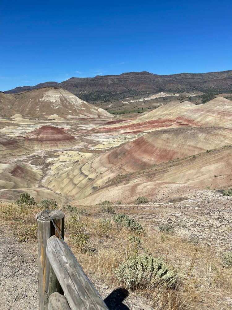 Painted Hills overlook - fence post in lower left quadrant and sloping rounded hills with bands of differently colored sediment of red, black, and yellow/brown. A vibrant blue sky spans above the hills. 