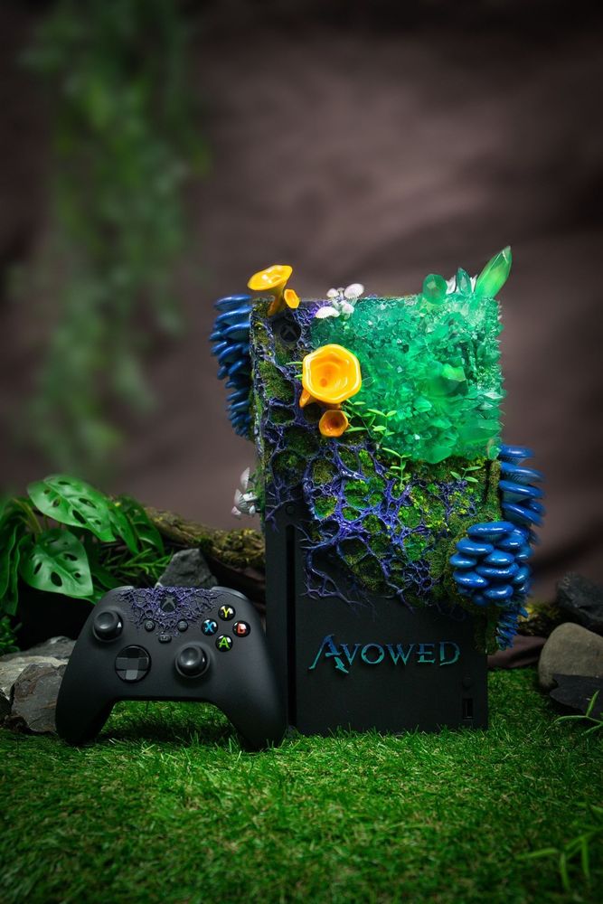 An Xbox Series X decorated with vibrant, nature-inspired elements including green crystals, blue lichen, and yellow flowers. The console is placed on a grassy surface with a matching controller in front of it. The word "AVOWED" is visible on the front of the console.(alt text generated using Microsoft Copilot and checked for accuracy)