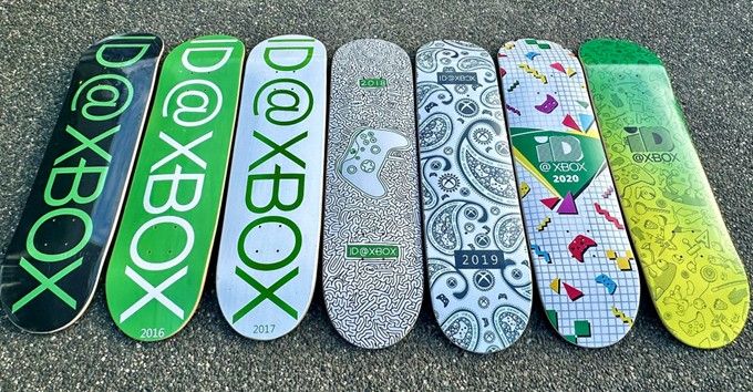 A row of seven skateboard decks laid out on a rough asphalt surface. Each deck features a unique design related to Xbox, with various years and patterns displayed. The designs include the Xbox logo, controllers, and abstract patterns in green, black, white, and other colors. (alt text generated using Microsoft Copilot and checked for accuracy)