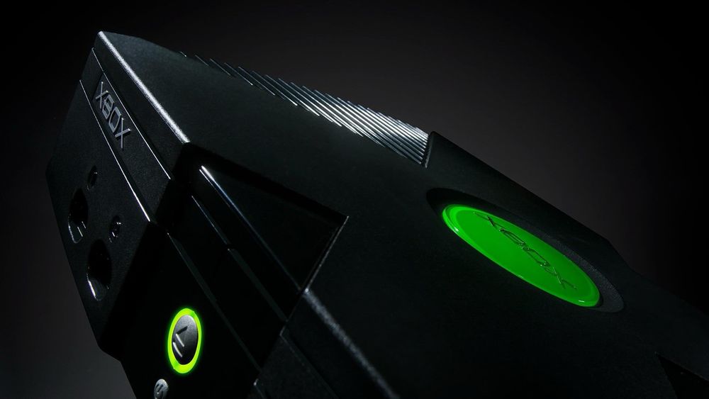 A close-up photo of an original Xbox console, featuring its black exterior and illuminated green power button. The iconic green circular Xbox logo is featured on the top, with a dark background.