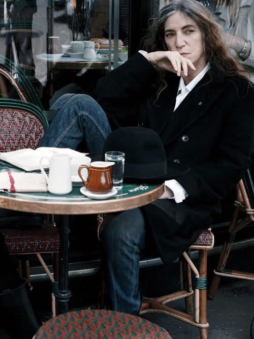 A young Patti Smith with long black hair at an outdoor table at a cafe 