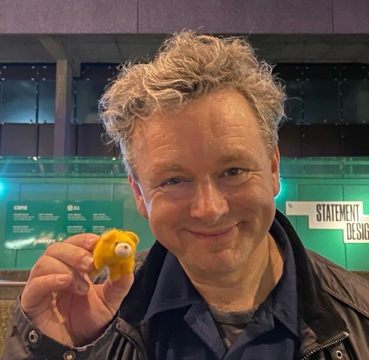 Michael Sheen smiling, while holding a small yellow (possibly) Bear. 