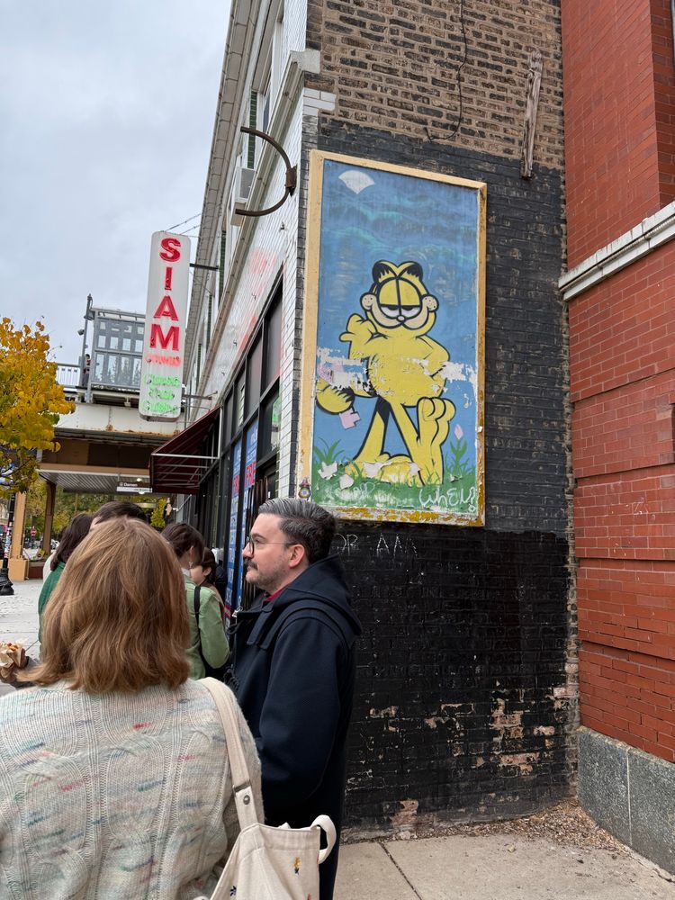 A long line of people outside of a bakery under the watchful eyes of Garfield painted on a brick wall 
