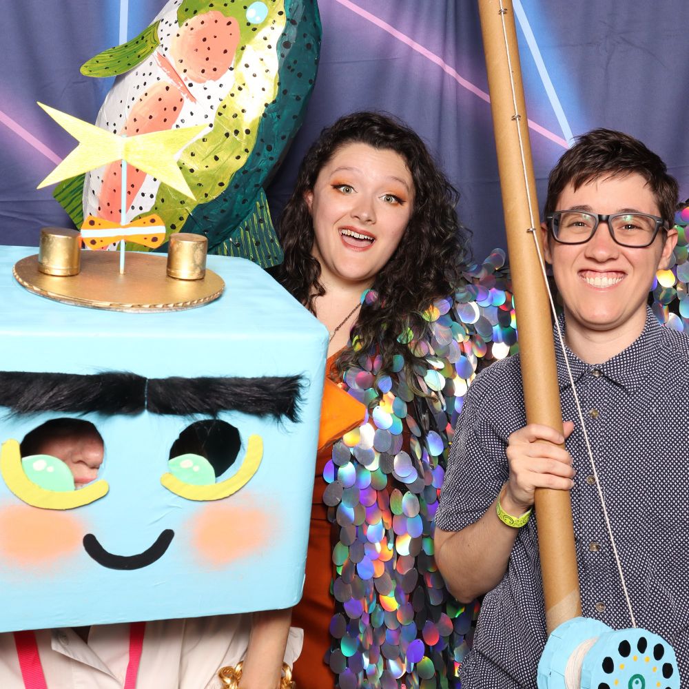 A Photo Booth photo of 3 people. One is wearing a robot head, I am wearing a sequin robe, and Coby is holding a comically large fishing pole with a paper mache fish on the end of its hook 