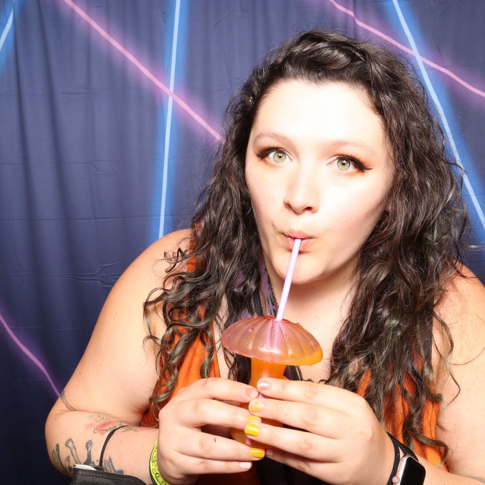 A photo booth photo of me sipping a beverage out of a mushroom shaped glass. I'm wearing an orange dress 
