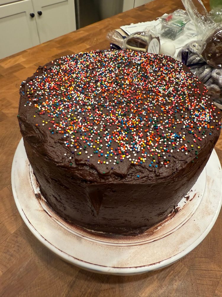 A top down photo of a poorly iced chocolate cake with a beautiful soul. It's topped with rainbow round sprinkles 