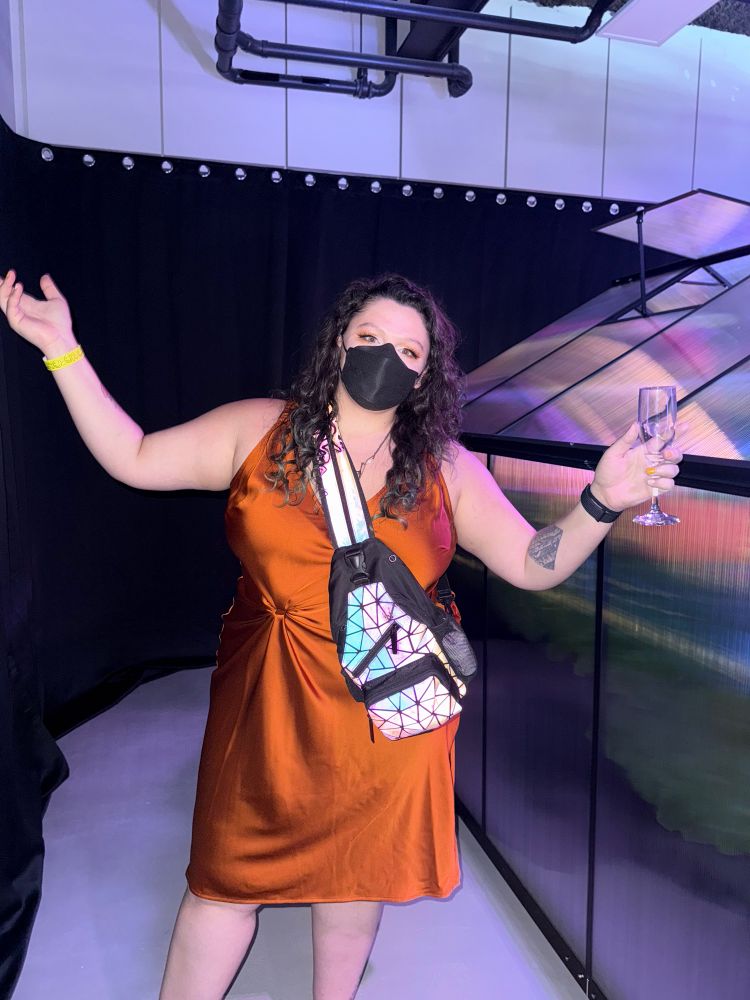 A photo of me in a satin orange dress holding a champagne flute and wearing a black mask. I am gesturing openly at the camera in a dark room 