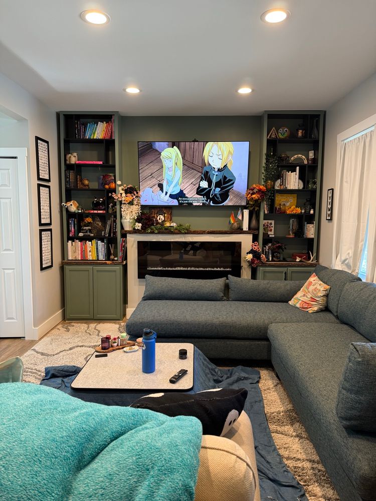 A photo of a living room with a mounted TV and a teal sectional couch. There are olive green cabinets that extend up into bookshelves with colorful trinkets and books reaching to the ceiling. A kotatsu is nestled between two couches 