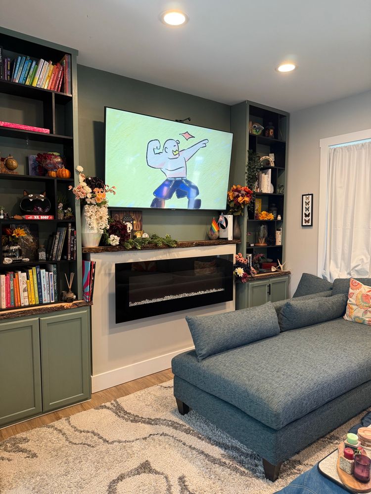 A photo of a living room with a mounted TV and a teal sectional couch. There are olive green cabinets that extend up into bookshelves with colorful trinkets and books reaching to the ceiling. Alex Louis Armstrong from Fullmetal Alchemist is flexing on the tv 