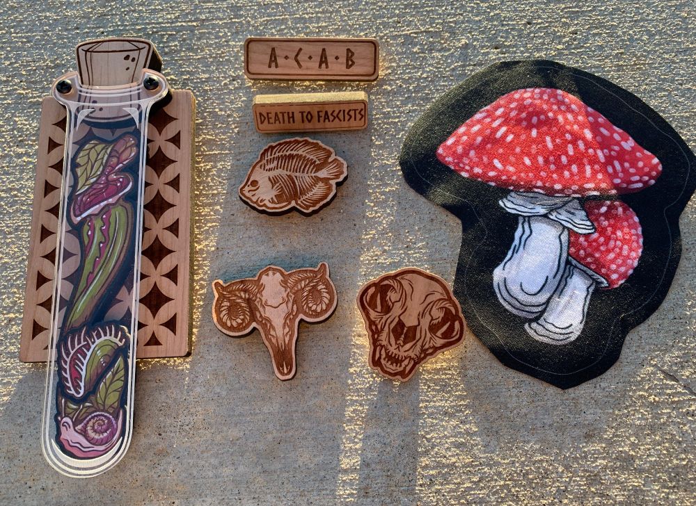 Laser engraved woodwork by Ghost of the Woods. On the left are carnivorous plants in a test tube, with a small snail at the bottom. In the middle a few wooden magnets and pins. On the right is a white-spotted mushroom with a bright red cap (Amanita muscaria) depicted on a black cloth.