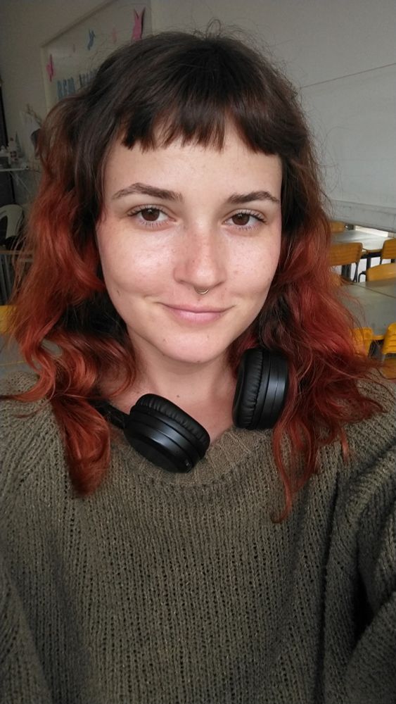 selfie de uma mulher branca de cabelos avermelhados (eu), usando fones de ouvido pendurados nos ombros, blusa de lã verde musgo e no fundo se vê uma sala de aula com cadeiras amarelas 