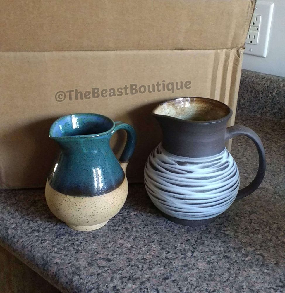 a photograph of two ceramic pitchers. On the left, the smaller pitcher is thrown from a tan, speckled body, with the upper half glazed in a deep blue-green. The pitcher on the right is made of a dark brown clay body, with a tan glaze on the inside, and white, ribbed slip on the outside. 

The shop name, the beast boutique, watermarks the image. 
