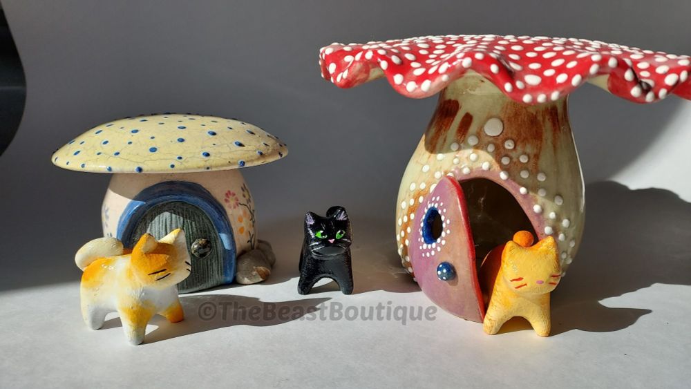 A photograph of two ceramic mushroom houses with three hand-painted cat figurines in front of them. 

On the left is a short white mushroom with a gray door and blue door frame. It's cap is yellow with small blue polka dots. Painted flowers run up the sides of the mushroom stem. In front of it is a white cat with orange splotches all over its body. 

Between the two mushrooms, facing the camera with its green eyes, is a black cat staring straight ahead. 
The mushroom on the right is taller with a purple door hanging ajar that has a blue door knob and a window in the middle. It's cap is red with scalloped edges and white polka dots. 

A ginger Tabby is peeking out of the door.

The shop name, the beast boutique, watermarks the image.