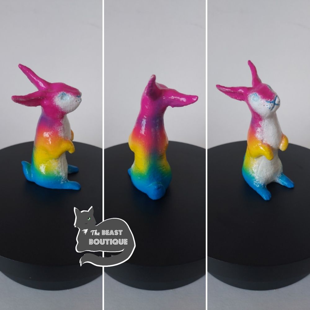 a collage showing a rabbit figurine standing in its hind legs from multiple angles. the rabbit is painted hot pink, yellow, and blue, like the colors of the pansexual flag, and has a white belly. its nose and mouth are blue.

the image is watermarked with a logo of a one-eyed grey cat, and the words "the beast boutique."