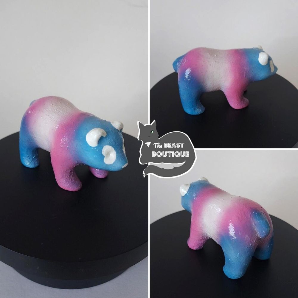 a collage showing a panda bear figurine from multiple angles. the panda is painted in the colors of the trans pride flag: blue, pink, and white. 

the image is watermarked with a logo of a one-eyed gray cat, and the words "the beast boutique."

