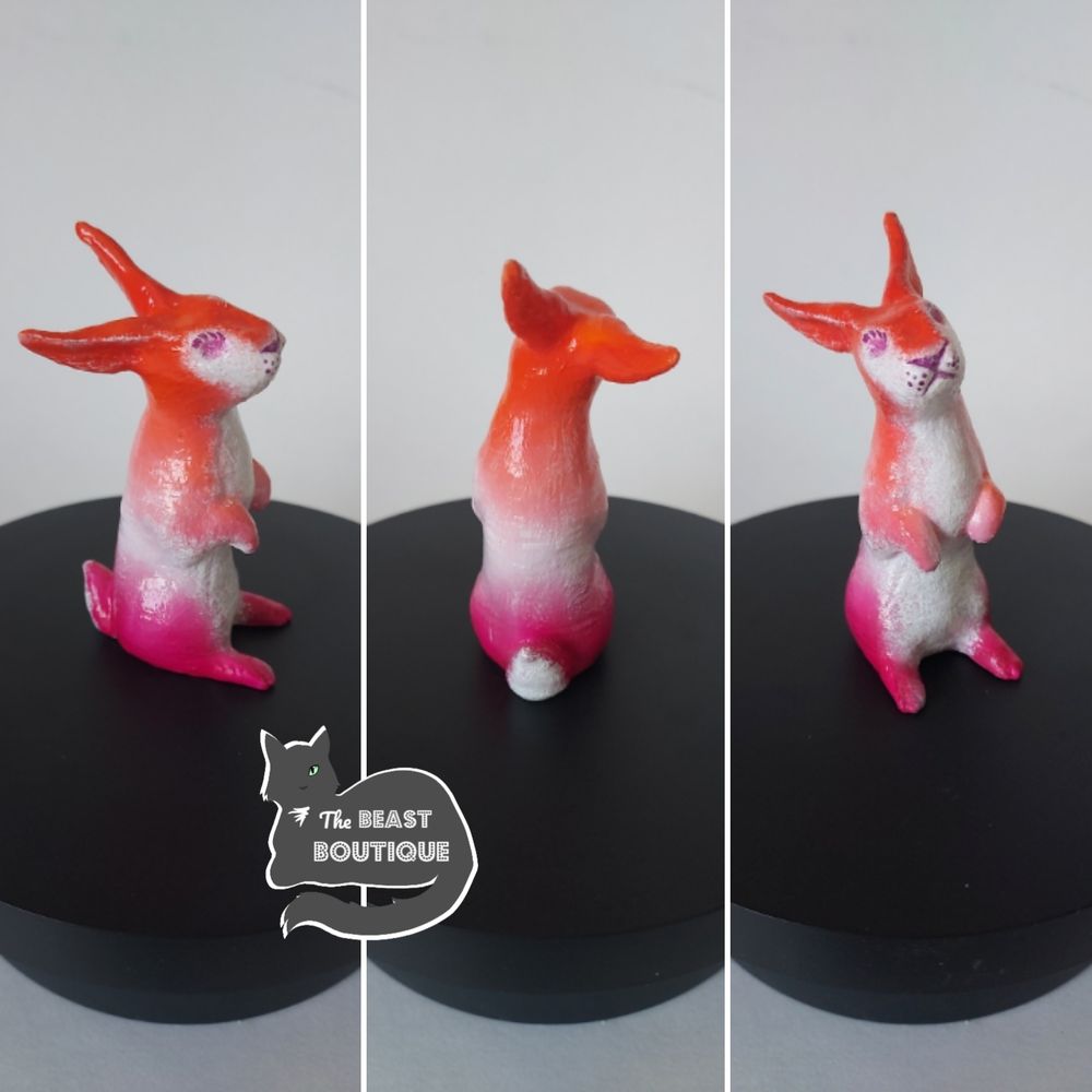a collage showing a rabbit figurine standing in its hind legs from multiple angles. the rabbit is painted orange, light orange, white, light pink, and magenta, like the colors of the lesbian flag, and has a white belly. its nose and mouth are magenta. 

the image is watermarked with a logo of a one-eyed grey cat, and the words "the beast boutique."