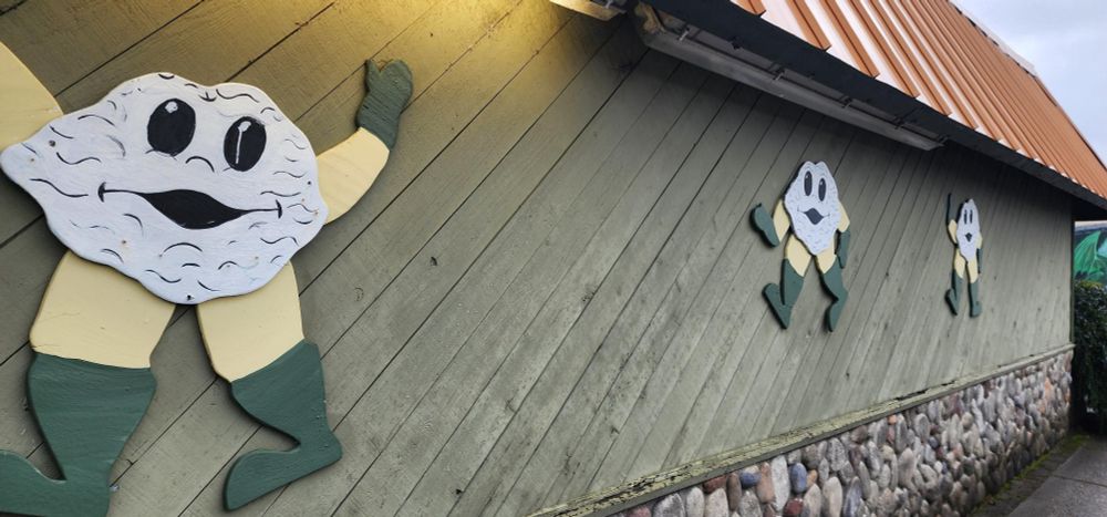 Picture of the wall of a low building called Shuckers where oysters run around with their dark soul less eyes, and giant thighs, wearing no pants and plotting something I am sure.  Translation, commercial art work cut outs of oysters with faces and arms and legs and gloves and boots but no pants.  These three oysters adorn a building, and look to be walking, waving, and raising their arms in victory.