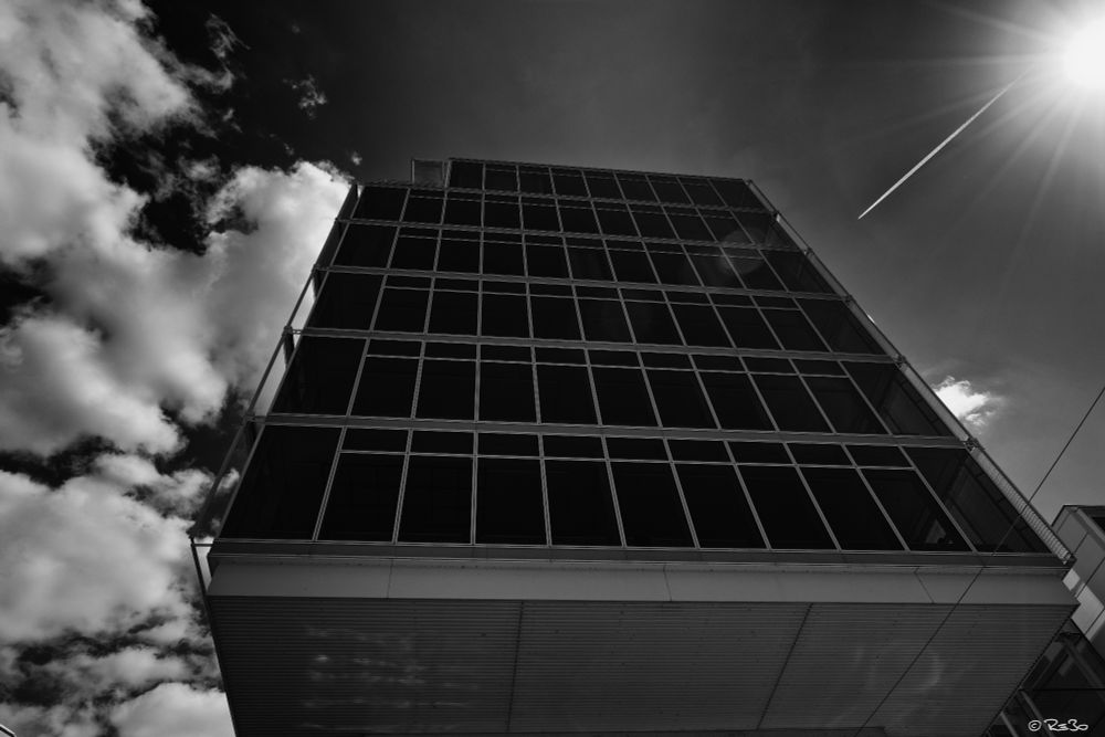 Black and white shot from below of a commercial building in front of a dark, cloudy sky, sun star in the upper right corner.