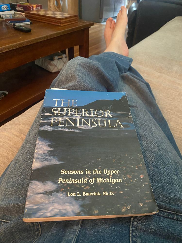 A picture of “The Superior Peninsula: Seasons in the Upper Peninsula of Michigan” by Lon L. Emerick on my lap with a wooden coffee table in the background. 