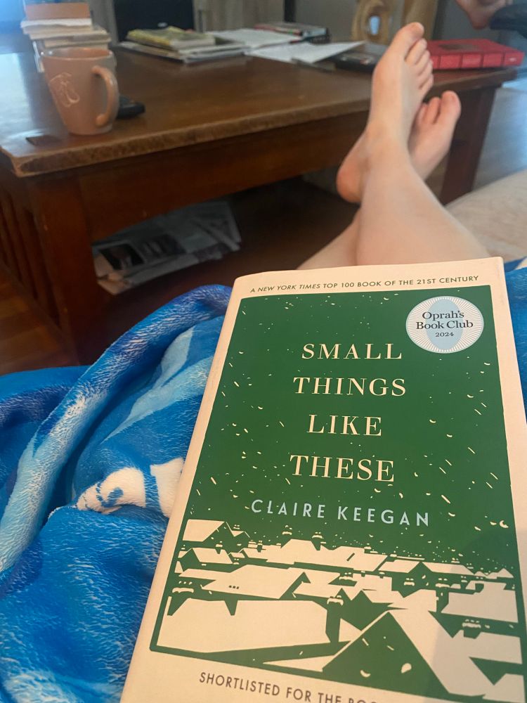 A green and white cover of Small Things Like These by Claire Keegan on my lap on the couch with a blue blanket, my bare feet and pink coffee cup in the background. 