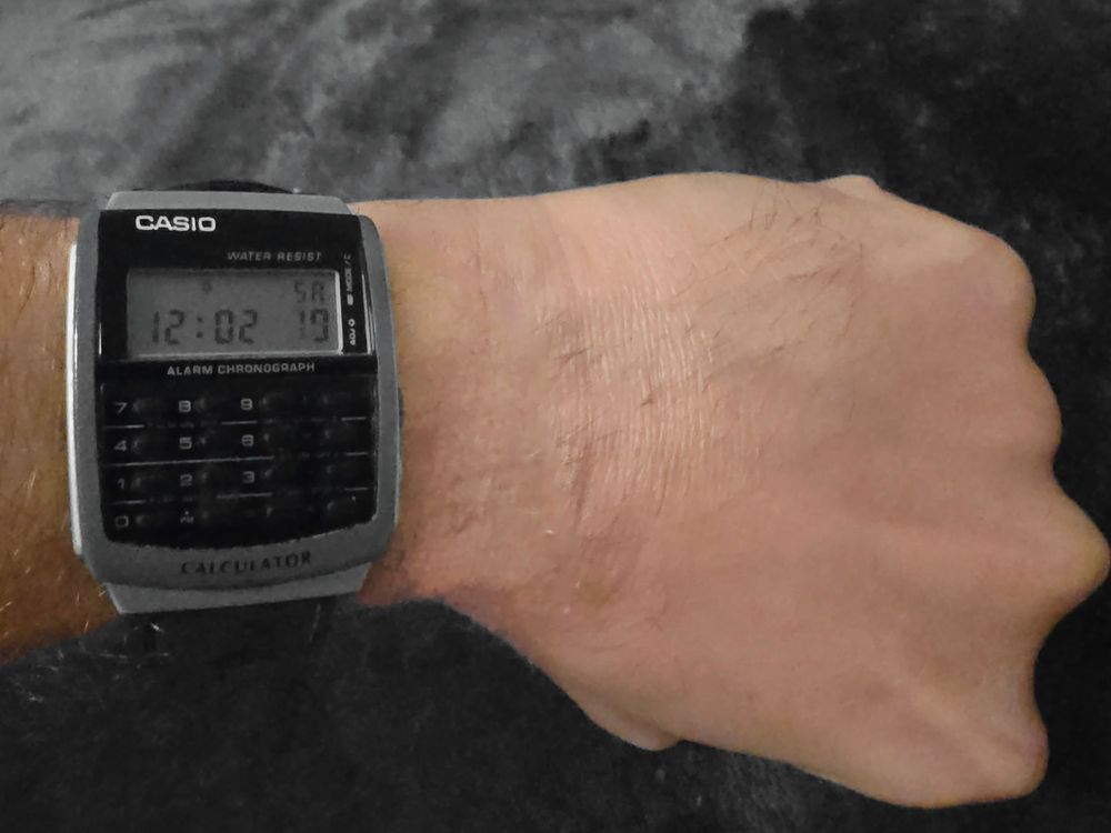 A Casio calculator watch worn on someone's wrist, showing the time as 12:02. The watch has a digital display and a calculator keypad below it. It's labeled as "WATER RESIST" and "ALARM CHRONOGRAPH" on the face. The watch has a silver/grey casing and is photographed against a dark background.