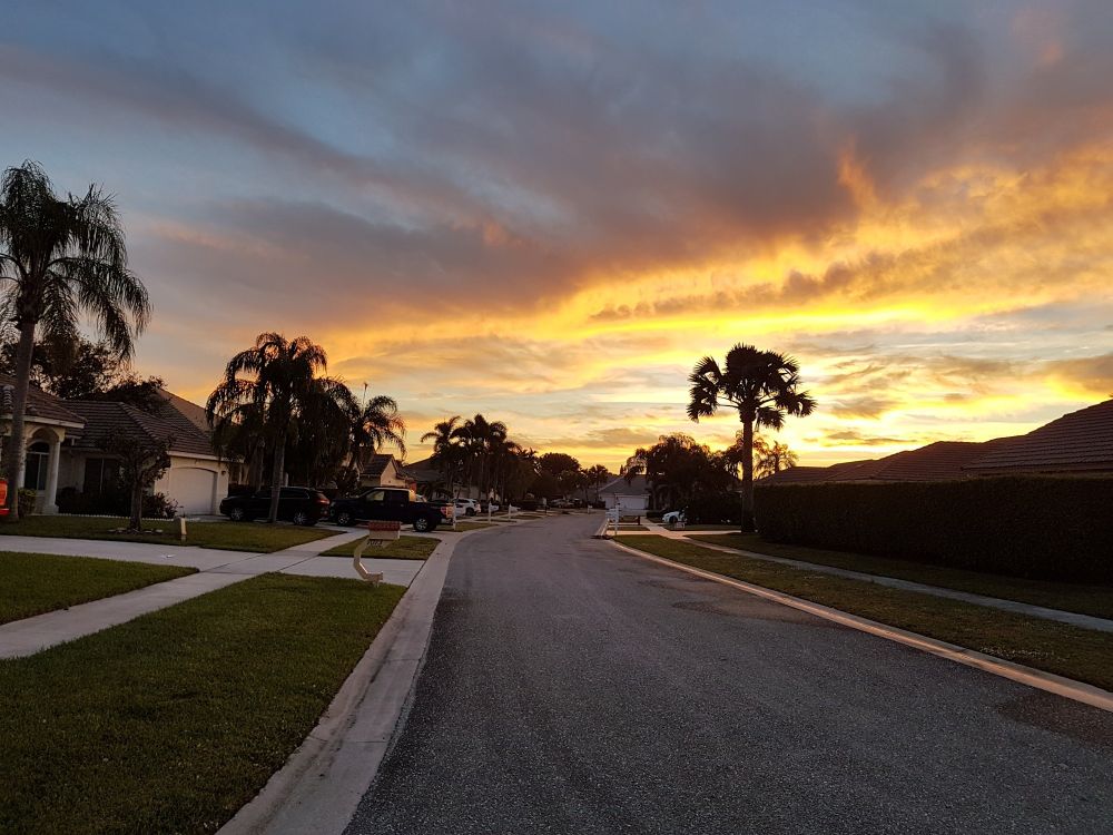A suburban street at sunset with palm trees silhouetted against a dramatic orange and yellow sky. Typical Florida single-story homes line the curved road.